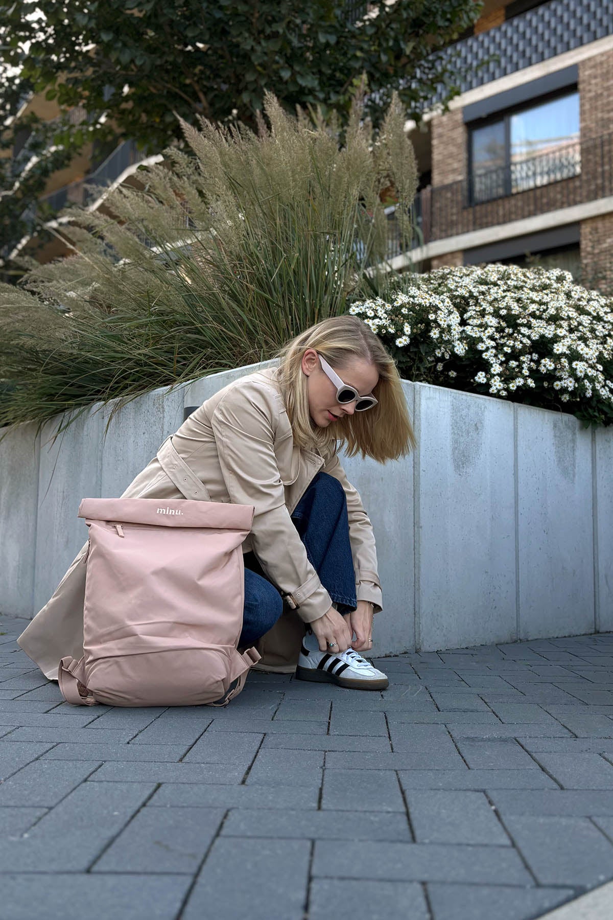 Woman tying her shoe next to a pink backpack on a paved area with plants and buildings in the background.